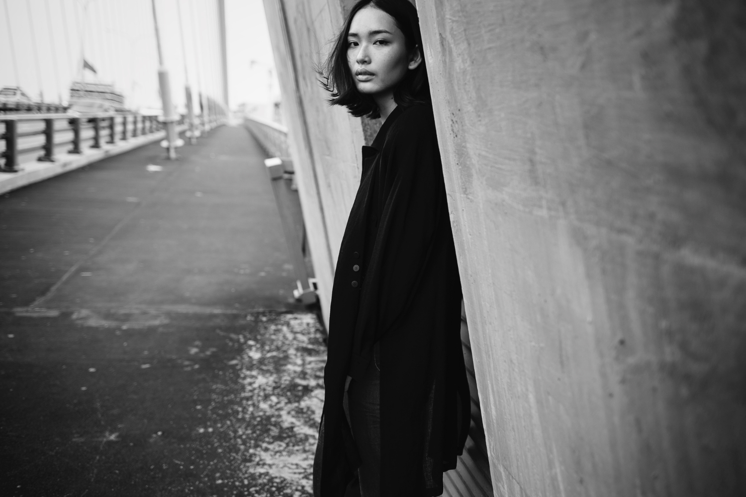 Black and white editorial portrait of a woman in a dark coat leaning against a concrete bridge span, looking back at the camera with quiet confidence - fashion as presence, not performance.